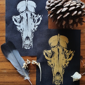 Op de afbeelding: Twee zwarte stoffen patches met een wit en gouden dieren schedel ontwerp. De schedels zijn gestileerd en hebben een gedetailleerd ontwerp. De patches bevinden zich op een houten oppervlak.