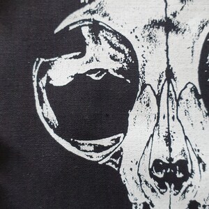May include: A white printed image of an animal skull on a black fabric background. The skull is facing the viewer with its mouth open and teeth visible.