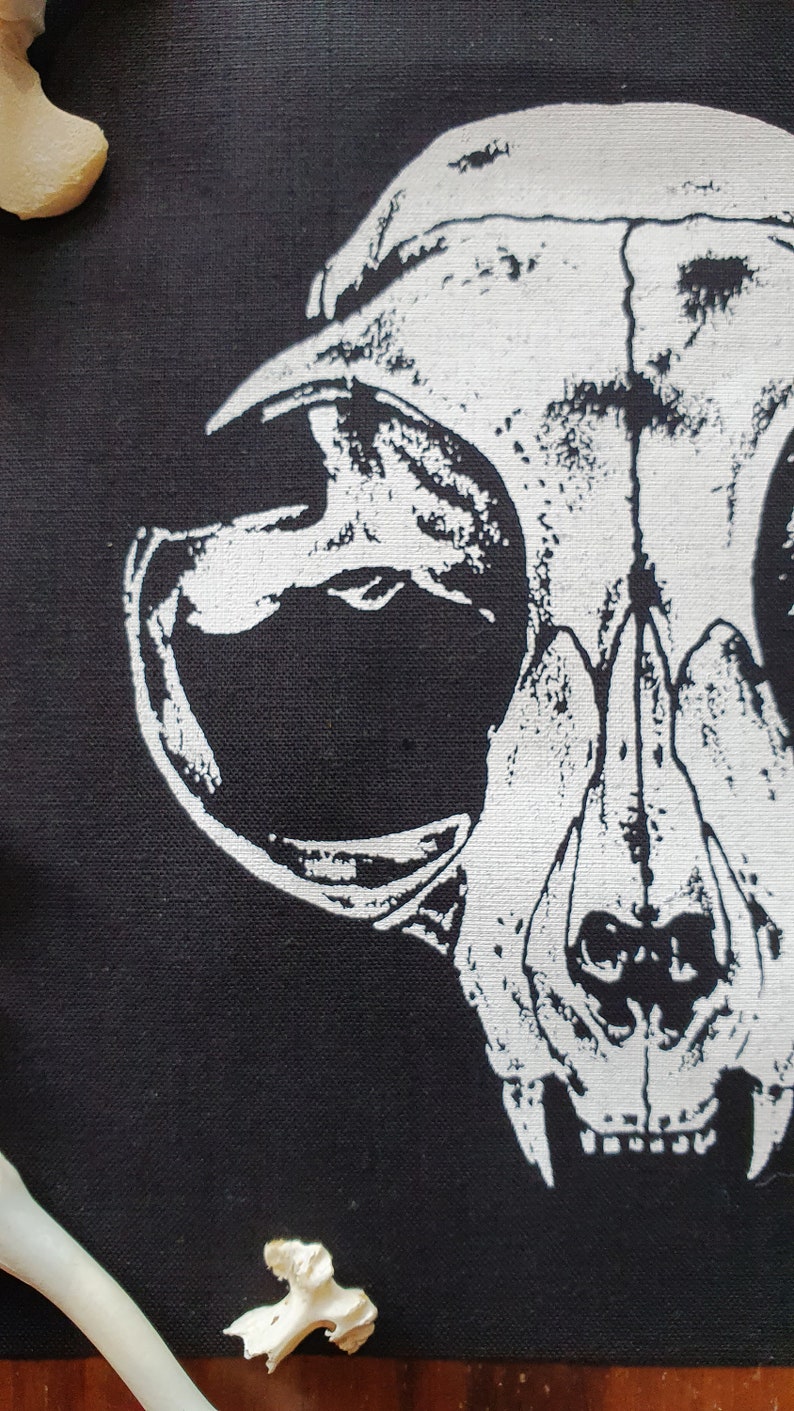 May include: A white printed image of an animal skull on a black fabric background. The skull is facing the viewer with its mouth open and teeth visible.
