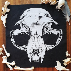 Peut inclure: Patch en tissu noir avec une image imprimée en blanc d'un crâne de chat. Le crâne est détaillé avec des dents et des orbites.