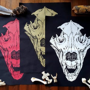 Puede incluir: Tres impresiones de tela negra con ilustraciones de cráneos de animales de diferentes colores. Los cráneos son blancos, rojos y amarillos. Las impresiones están en tela negra y tienen un aspecto desgastado.
