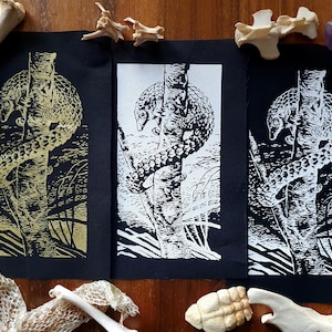 Könnte beinhalten: Drei schwarze Stoffdrucke mit einem weißen Schuppentier, das an einem Baum hochklettert. Die Drucke werden auf einer Holzoberfläche mit Knochen und anderen natürlichen Objekten gezeigt.