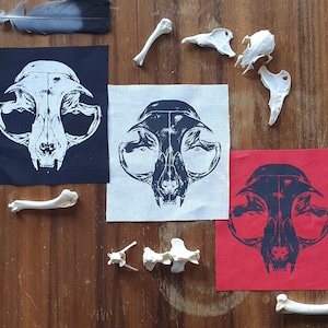 Könnte beinhalten: Drei Stoffpatches mit einem schwarz-weißen Bild eines Katzen Schädels darauf gedruckt. Die Patches liegen auf einer Holzoberfläche mit weißen Tierknochen, die um sie herum verstreut sind. Die Patches sind schwarz, weiß und rot.