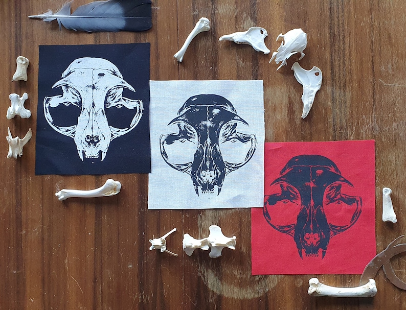 May include: Three fabric patches with a black and white image of a cat skull printed on them. The patches are on a wooden surface with white animal bones scattered around them. The patches are black, white, and red.
