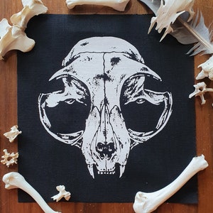 May include: Black fabric patch with a white printed image of a cat skull. The skull is detailed with eye sockets, a nose, and teeth. The patch is surrounded by white bones.