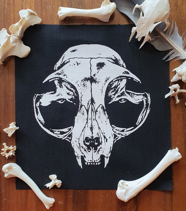 May include: Black fabric patch with a white printed image of a cat skull. The skull is detailed with eye sockets, a nose, and teeth. The patch is surrounded by white bones.
