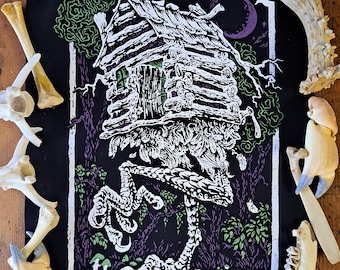 May include: A black fabric patch featuring a white, green, and purple screen-printed illustration of a log cabin standing on giant bird legs. The design is surrounded by a white rectangular border and various animal bones and a seashell.