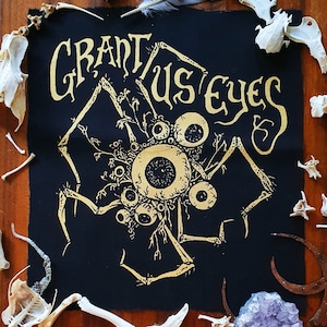 Op de afbeelding: Zwarte stoffen patch met gouden borduurwerk van een wezen met meerdere ogen en dunne poten en de tekst "Grant Us Eyes".