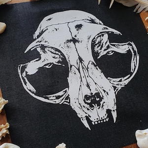 May include: A black fabric patch with a white printed image of a cat skull. The skull is detailed with teeth and eye sockets. The patch is surrounded by white bones on a wooden surface.