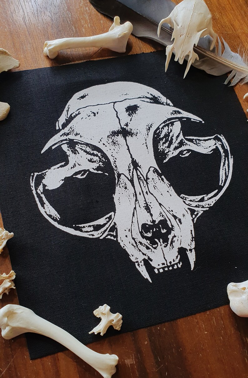 May include: A black fabric patch with a white printed image of a cat skull. The skull is detailed with teeth and eye sockets. The patch is surrounded by white bones on a wooden surface.