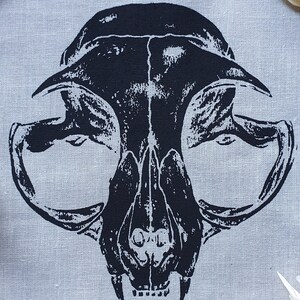 May include: Black and white printed fabric patch featuring a cat skull design. The skull is detailed with teeth and eye sockets. The patch is on a light gray fabric background.