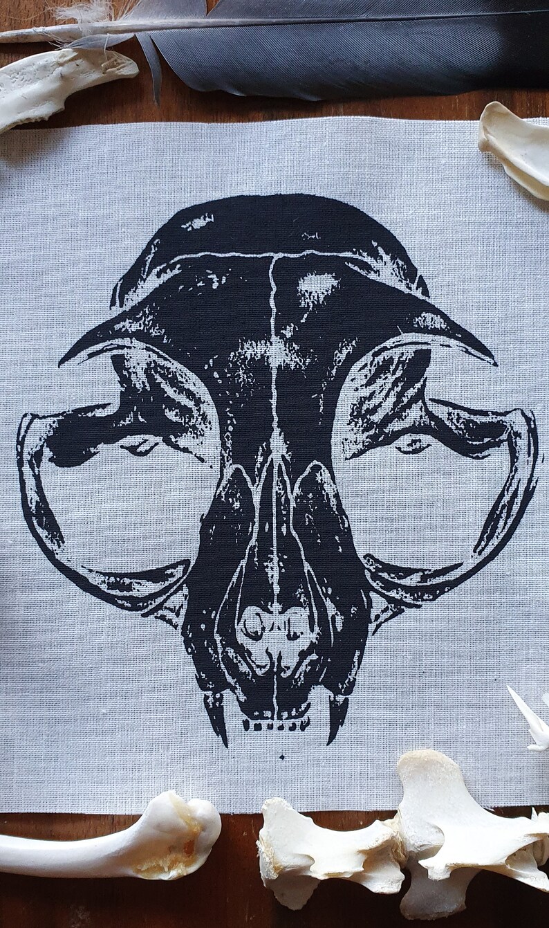 May include: Black and white printed fabric patch featuring a cat skull design. The skull is detailed with teeth and eye sockets. The patch is on a light gray fabric background.
