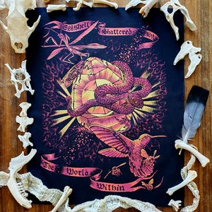 Könnte beinhalten: Schwarzer Kunstdruck mit einer farbenfrohen Illustration einer Schlange, eines Kolibris und einer Gottesanbeterin. Der Text lautet "Eggshell Shattered By The World Within". Umgeben von Knochen und einer Feder auf Holz.