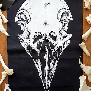 Puede incluir: Un parche de tela negro con una imagen impresa en blanco de una calavera de pájaro con un pico largo. La calavera es detallada y realista.