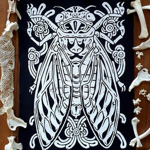 May include: A black and white illustration of a cicada with intricate details. The cicada is depicted with large wings and a detailed head and body. The illustration is printed on a black fabric.