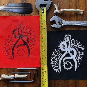 May include: Two iron-on patches with a black and white design of a pair of pliers with a worm wrapped around them. The text "PROCESS" is written around the pliers. The patches are on a red and black fabric background.