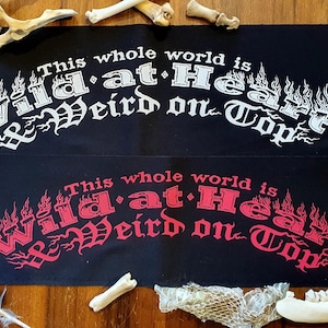 Op de afbeelding: Twee zwarte stoffen patches met de tekst "This whole world is Wild at Heart & Weird on Top" in witte en rode letters. De bovenste patch heeft witte letters en de onderste patch heeft rode letters.