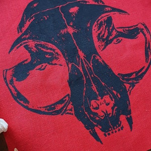 May include: Black and white skull design printed on a red fabric patch. The patch is surrounded by white bones.