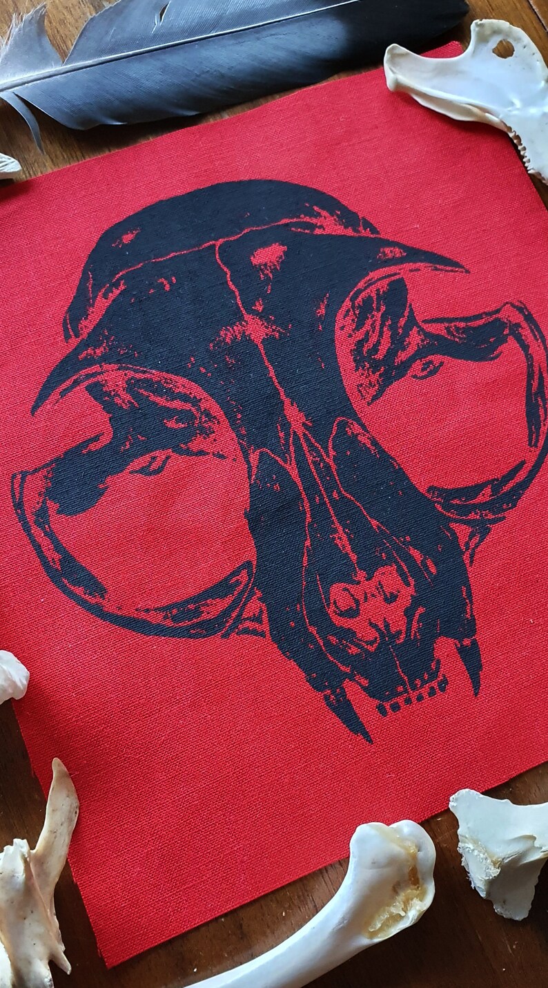 May include: Black and white skull design printed on a red fabric patch. The patch is surrounded by white bones.