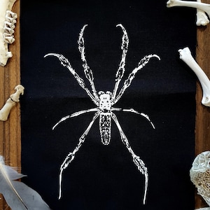Könnte beinhalten: Schwarzer Stoff-Patch mit einer weißen Silhouette einer Spinne mit langen Beinen. Die Spinne ist mittig auf dem Patch platziert.