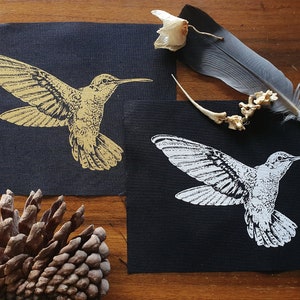 Könnte beinhalten: Zwei schwarze Stoffpatches mit einem Kolibri-Design. Ein Patch zeigt einen goldenen Kolibri, der andere Patch zeigt einen weißen Kolibri.