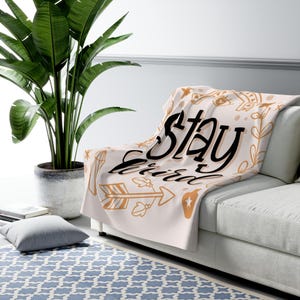 May include: A cream-colored throw blanket with the words "Stay Weird" in black script. The blanket features gold-colored illustrations of arrows, stars, and floral elements. The blanket is draped over a white sofa.