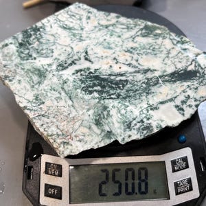 May include: A slab of moss agate, displaying a mix of white and green patterns, rests on a digital scale. The scale shows a weight of 250.8 grams. The agate's surface has intricate, natural designs.