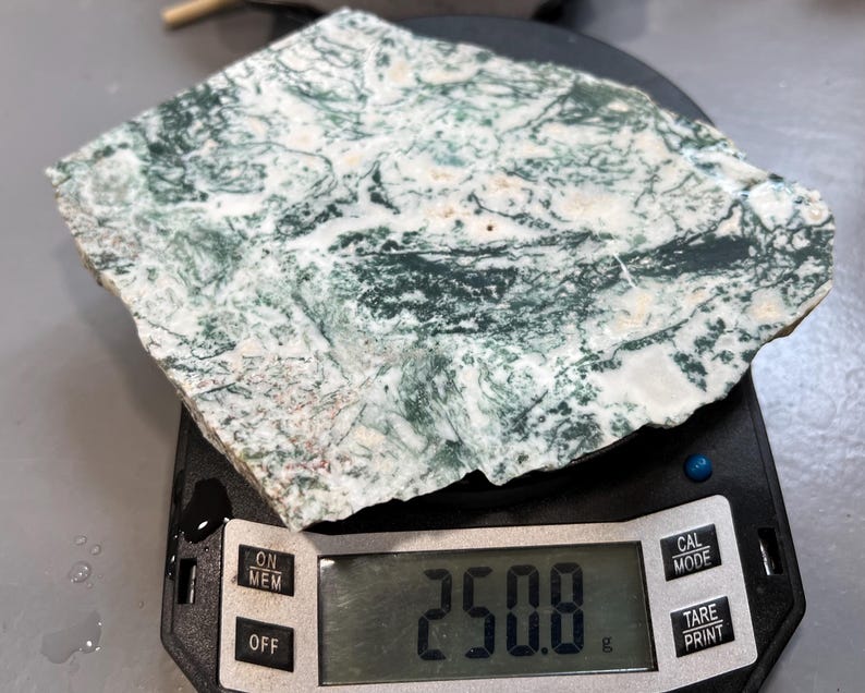 May include: A slab of moss agate, displaying a mix of white and green patterns, rests on a digital scale. The scale shows a weight of 250.8 grams. The agate's surface has intricate, natural designs.