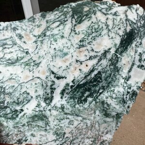 May include: A rough-cut slab of moss agate stone, showcasing a natural pattern of green, white, and tan hues. The stone's surface displays intricate, vein-like formations, creating a unique and organic aesthetic. The stone is held in a hand.
