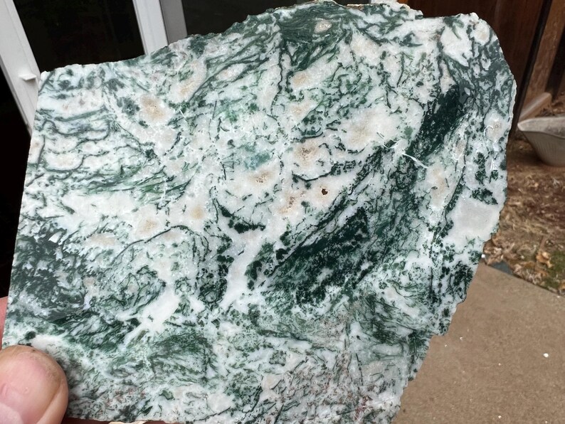 May include: A rough-cut slab of moss agate stone, showcasing a natural pattern of green, white, and tan hues. The stone's surface displays intricate, vein-like formations, creating a unique and organic aesthetic. The stone is held in a hand.