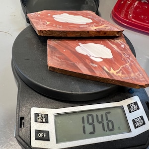 May include: Two reddish-brown, square-shaped wood pieces with white and yellow accents are placed on a digital scale. The scale displays a weight of 194.6 grams. The wood pieces have a unique grain pattern.