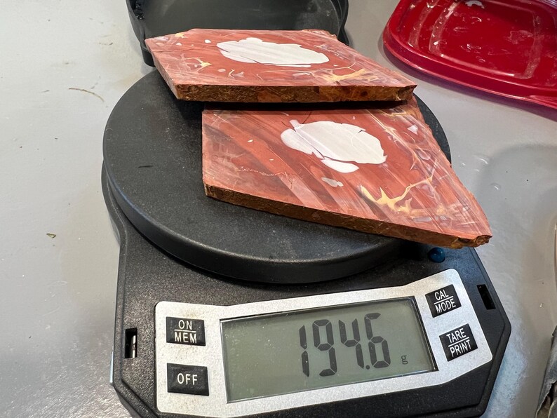 May include: Two reddish-brown, square-shaped wood pieces with white and yellow accents are placed on a digital scale. The scale displays a weight of 194.6 grams. The wood pieces have a unique grain pattern.