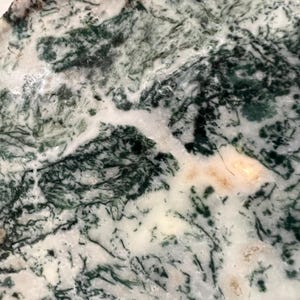 May include: Close-up of a polished stone with a natural pattern. The stone features a mix of white, dark green, and light brown colors, creating a marbled effect. The intricate veining and color variations give the stone a unique appearance.