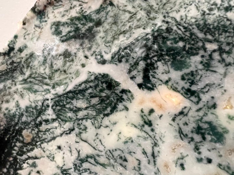 May include: Close-up of a polished stone with a natural pattern. The stone features a mix of white, dark green, and light brown colors, creating a marbled effect. The intricate veining and color variations give the stone a unique appearance.