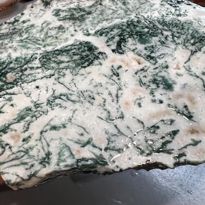May include: A natural stone slab with a unique pattern. The stone features a white base with intricate green and brown veining, creating a marbled effect. The surface has a slightly wet appearance, enhancing the colors and details of the stone.