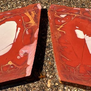May include: Two polished, irregularly shaped stones. The stones are primarily red with white and gold veining and patterns. The stones have a natural, organic appearance, with visible cracks and variations in color. The overall aesthetic is earthy and unique.