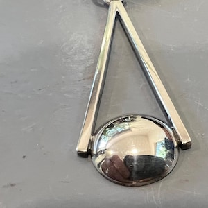 May include: A silver-toned pendant necklace featuring a geometric design. The pendant has a polished, spherical base and a triangular frame. The necklace is displayed on a gray surface.