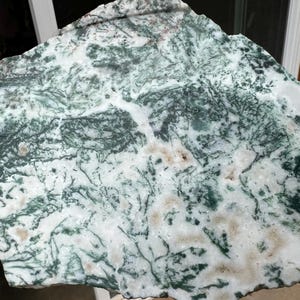 May include: A natural stone slab with a unique pattern of white, green, and brown hues. The stone's surface features intricate, vein-like patterns, creating a textured appearance. The irregular shape and color variations suggest a raw, organic origin.