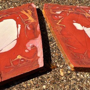 May include: Two rectangular, polished stone slabs. The slabs are primarily red with white and gold veining. The stone has a natural, organic appearance, with variations in color and texture. The slabs are displayed on a textured, light brown surface.