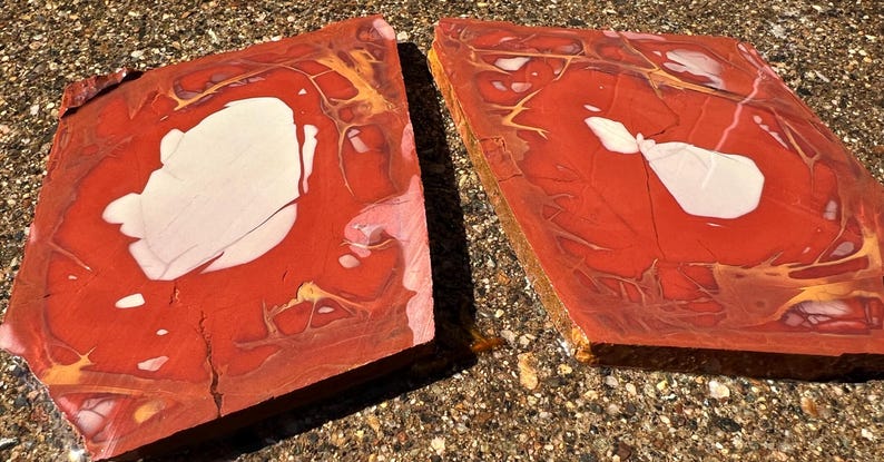 May include: Two rectangular, polished stone slabs. The slabs are primarily red with white and gold veining. The stone has a natural, organic appearance, with variations in color and texture. The slabs are displayed on a textured, light brown surface.