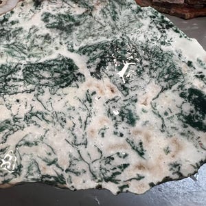 May include: A natural stone slab with a unique pattern of green, white, and tan colors. The stone has a polished surface with a network of dark green veins and patterns resembling a landscape. This decorative stone is ideal for home decor.