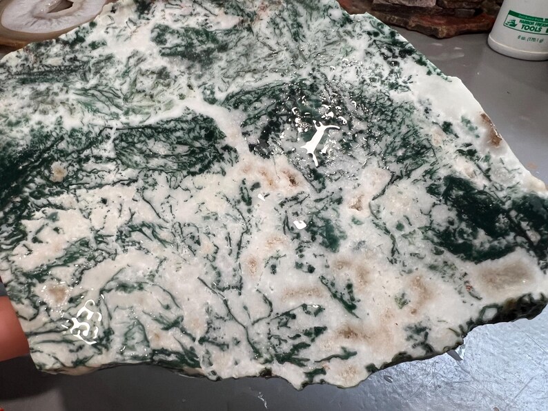 May include: A natural stone slab with a unique pattern of green, white, and tan colors. The stone has a polished surface with a network of dark green veins and patterns resembling a landscape. This decorative stone is ideal for home decor.