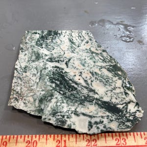 May include: A rough-cut stone slab with a marbled pattern of white and dark green. The stone is irregularly shaped and lies on a gray surface. A wooden ruler with inch markings is at the bottom.