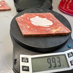 May include: A red and white patterned wooden tile is placed on a digital scale, which reads 99.3 grams. The tile has a white central design and is surrounded by red and yellow hues. The scale is black.