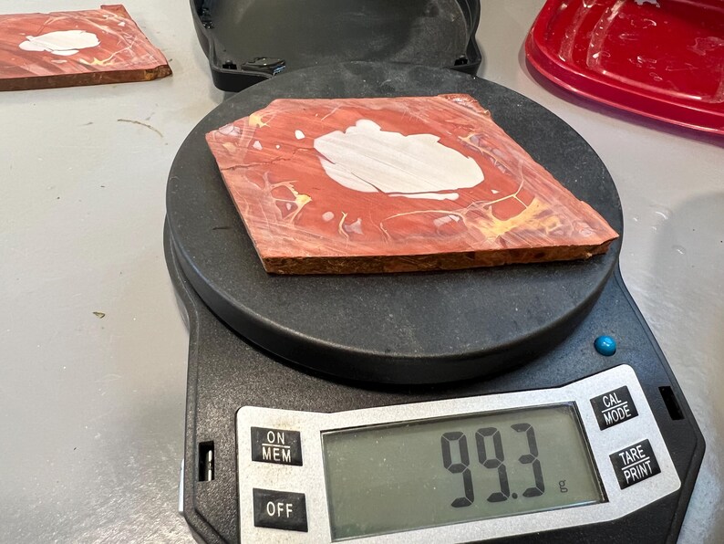 May include: A red and white patterned wooden tile is placed on a digital scale, which reads 99.3 grams. The tile has a white central design and is surrounded by red and yellow hues. The scale is black.