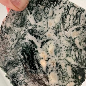May include: A close-up of a polished stone slab with a natural pattern. The stone features a mix of white, cream, and dark green colors, creating a marbled effect. The edges are rough, and the overall shape is irregular.