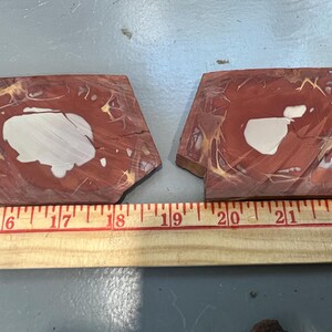 May include: Two pieces of polished red jasper stone with white and tan patterns. The stones are irregularly shaped and measure approximately 5 to 6 inches in length. A wooden ruler is placed below the stones.