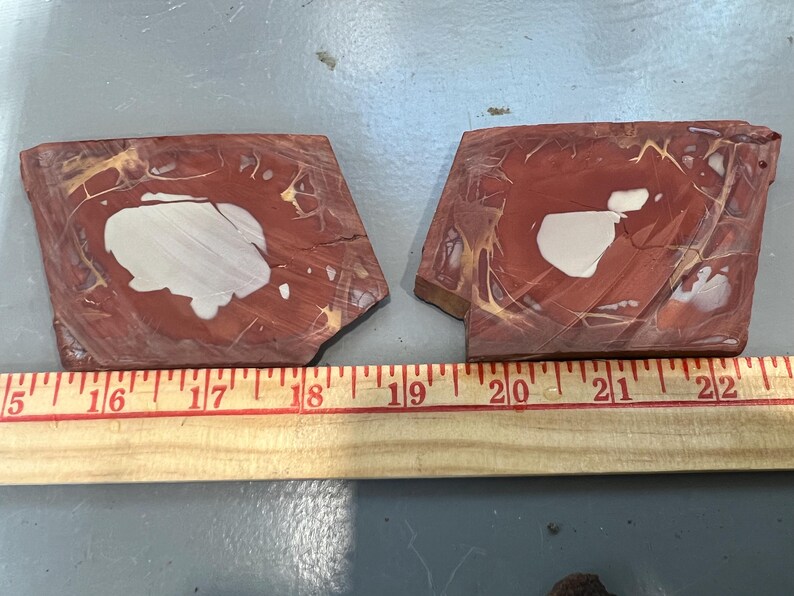 May include: Two pieces of polished red jasper stone with white and tan patterns. The stones are irregularly shaped and measure approximately 5 to 6 inches in length. A wooden ruler is placed below the stones.