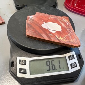 May include: A piece of red and white patterned stone sits on a digital scale, displaying a weight of 96.1 grams. The scale is black, with buttons for power and other functions. Another piece of stone is visible to the left.