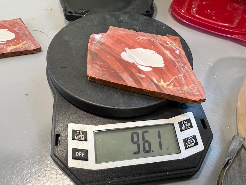 May include: A piece of red and white patterned stone sits on a digital scale, displaying a weight of 96.1 grams. The scale is black, with buttons for power and other functions. Another piece of stone is visible to the left.